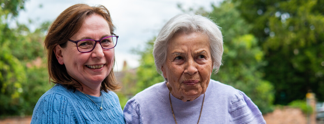 Eine Frau mittleren Alters mit Brille stützt eine Senorin, die eine Halskette trägt, beim Gehen. Im Hintergrund sind grüne Bäume.