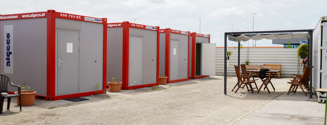 Mehrere rot-graue Container im Freien, gegenüber ein überdachter Sitzbereich.